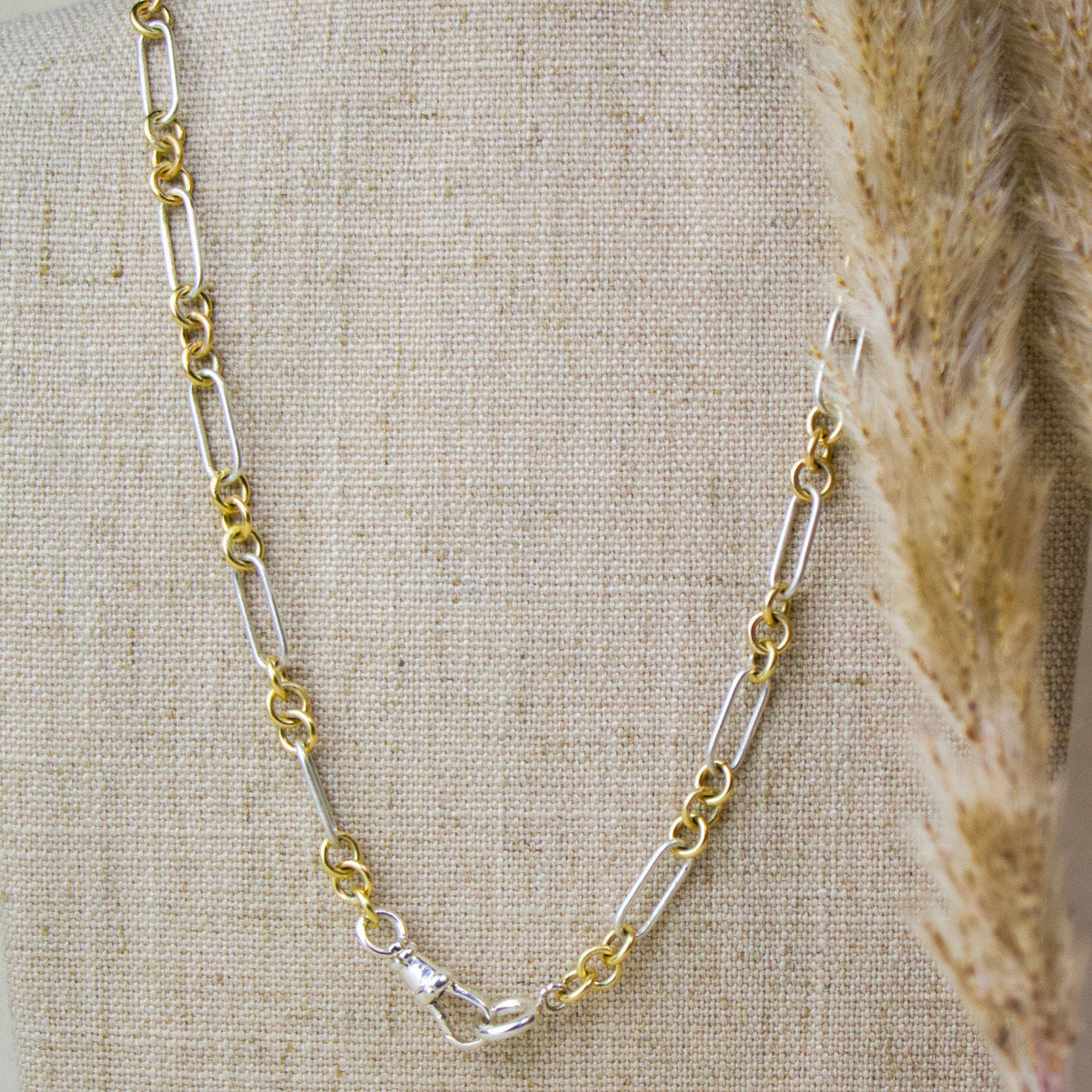 A two-tone chain necklace with alternating gold and silver links, displayed on a beige linen jewelry bust with dried pampas grass on the right side.