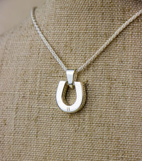 Back View of a silver horseshoe pendant necklace on a beige fabric background. Hand engraved H on the back