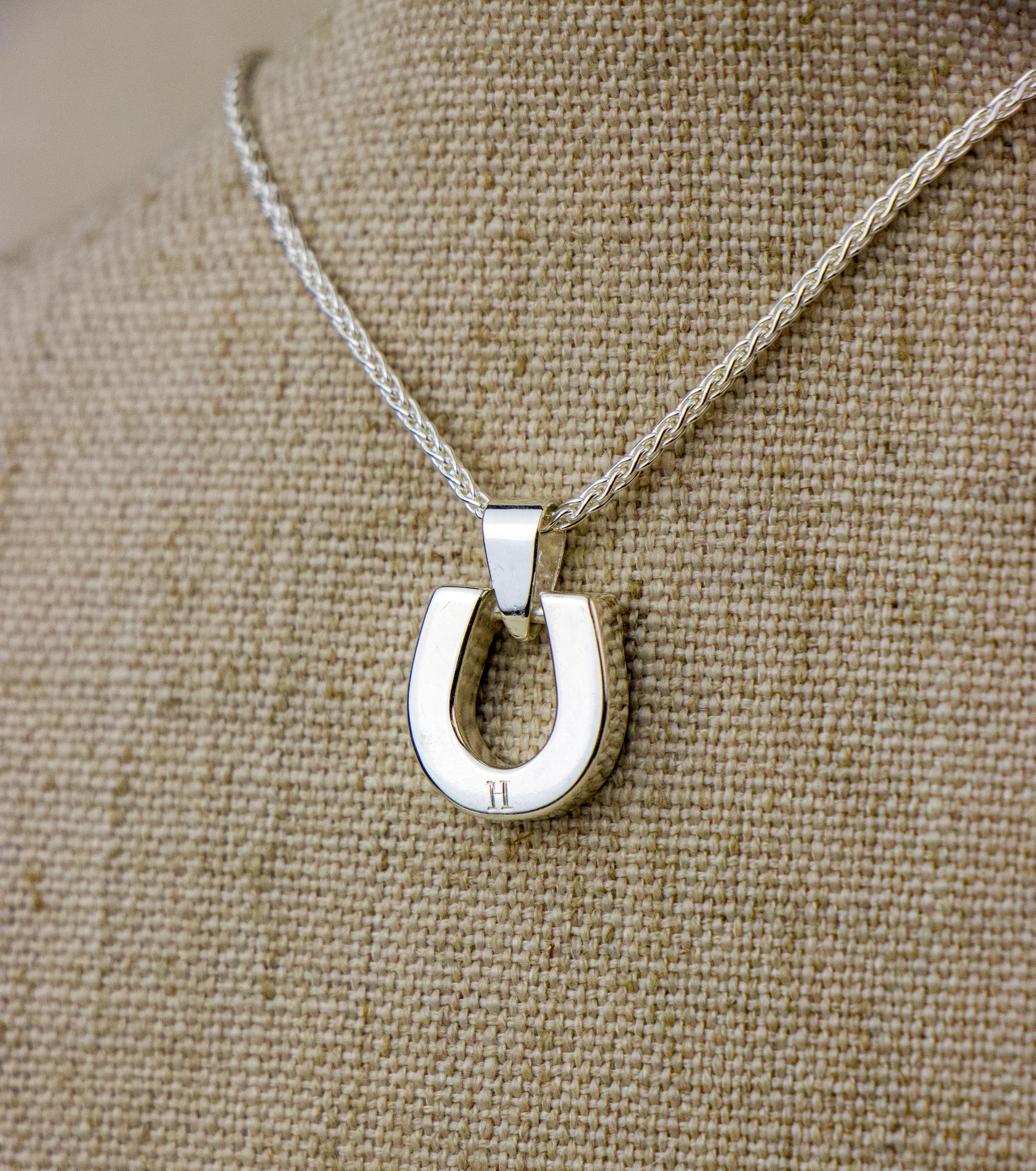 Back View of a silver horseshoe pendant necklace on a beige fabric background. Hand engraved H on the back