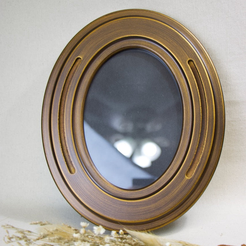 An oval picture frame with a textured bronze-gold metallic frame and a dark reflective glass center, styled with dried grasses in the foreground against a neutral light gray background.