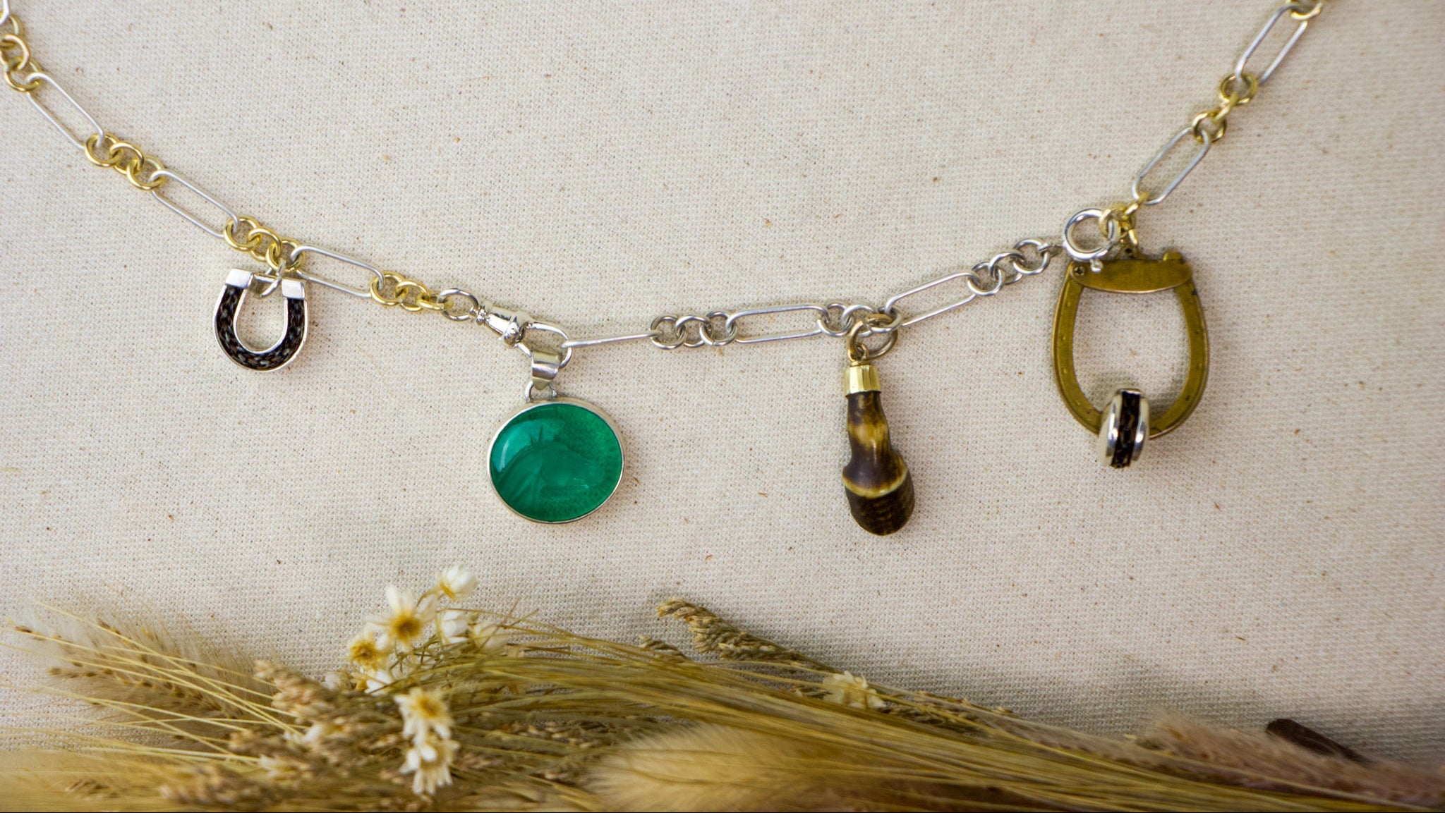 A silver and gold chain necklace with four unique charms: a horseshoe, a round green cabochon, a small tortoiseshell bead, and a gold horse bit, displayed on a textured beige fabric background with dried wheat.