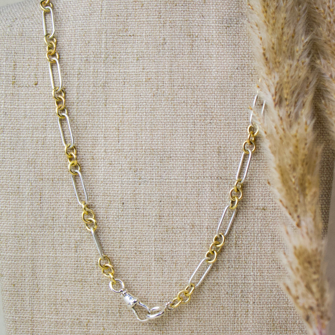 A two-tone chain necklace with alternating gold and silver links, displayed on a beige linen jewelry bust with dried pampas grass on the right side.