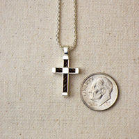 A silver cross pendant with horsehair inlay, shown on a silver chain next to a US dime for scale, on a beige textured fabric background.