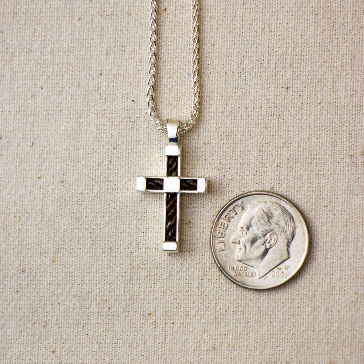 A silver cross pendant with horsehair inlay, shown on a silver chain next to a US dime for scale, on a beige textured fabric background.