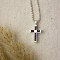 A silver cross pendant necklace with a horsehair inlay, hanging from a silver chain against a beige linen background with dried wheat stalks in the foreground.